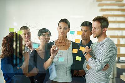 Group of people crowded around a glass wall, discussing the writing and post-it notes that are on the glass