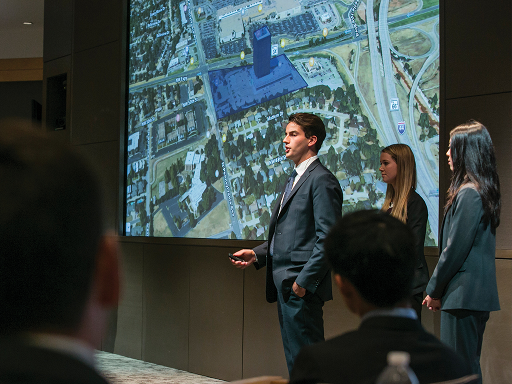 Three people in business attire stand in front of a projector screen presenting to a group.
